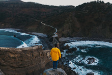 From Above Back View Of Faceless Man In Vivid Yellow Jacket And Denim Standing On Edge Of Cliff And Looking At Empty Road Crossing Old Stone Bridge Among Boulders And Troubled Waves Of Bay To Forested Hills In Spain