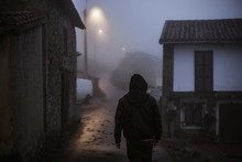 Back View Of Faceless Male In Hood Walking On Rural Road At Night In Village