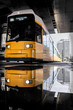 yellow tram is reflected in a street puddle on alexander Platz in the centre of Berlin, the train goes through a dark bridge, in the background there is a white blurred high-rise building