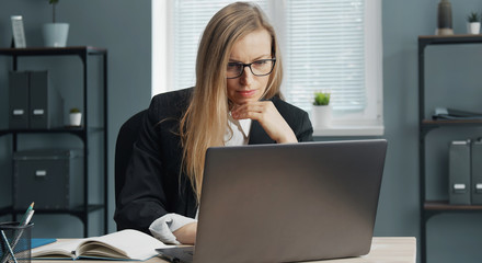 Focused mature business lady in formal suit and specs working on laptop sitting at office table