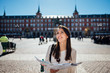 © eldarnurkovic - Visiting famous landmarks and places.Cheerful female traveler at famous Plaza Mayor square reading a map. Marid,Spain travel experience. Backpacker, travel photography.
