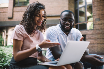 Cheerful dark skinned male and female friends making video call together via laptop computer and accessory outdoors, positive african american couple satisfied with 4G connection watching video