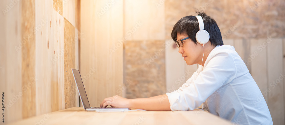 Young Asian man wearing headphones listening to music while working with laptop computer. Male office worker doing online conference with his business company team. Work from home concept - obrazy, fototapety, plakaty 