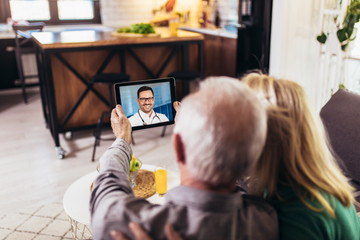 Senior couple at home holding digital tablet during video call with family doctor