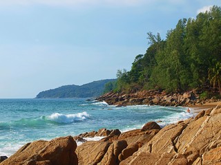 Panoramic view of rocky and sandy shore of a wild beach. Landscape of sea bay, the azure water and the crest of a wave rolling to the coast. Hills covered with green forest at the horizon.