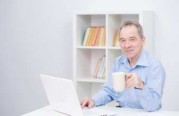 Handsome elderly man working on laptop, smiling, looking at camera, drinking coffe.