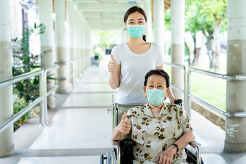 Young Asian grandchild taking care her grandmother sitting on wheelchair. Grandmother almost 90 years old was take care by her granddaughter while traveling at park. People wearing protective mask.