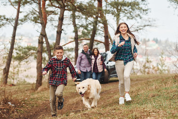 Happy family runs and having fun with their dog near modern car outdoors in forest