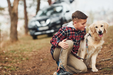 Wall Mural - Cheerful boy in casual clothes sitting with her dog in forest against modern black car