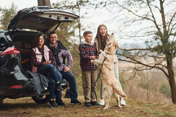 Wall Mural - Happy family have fun with their dog near modern car outdoors in forest