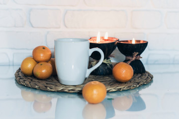 Wall Mural - Burning spa aroma candles in coconut shell, cup of tea, tangerines on wicker stand on a glass white table, cozy home interior background.