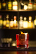 Red cocktail with ice decorated with orange peel on a bar counter against the background of alcohol bottles.