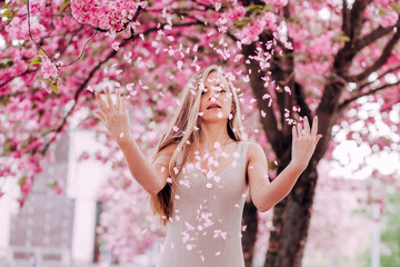 Wall Mural - Closeup portrait of a lovely girl in a park with blooming japanese sakura trees. Romantic young blonde in a dress posing on a background of spring flowering trees. Flying cherry petals.