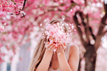 Wall Mural - Closeup portrait of a lovely girl in a park with blooming japanese sakura trees. Romantic young blonde in a dress posing on a background of spring flowering trees. Flying cherry petals.