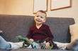 © Strelciuc - Caucasian small boy sitting on the sofa and playing with his dinosaur toys while looking at his parents