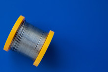 Silver Threads Of Solder Are Wound On A Yellow Spool. S. The Coil With Solder Lies On A Dark Blue Background.