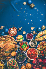 Roasted Christmas turkey with orange slices, cranberries, garlic, festive decoration, candles, tangerine, pomegranate, golden cultery, star glitter sparkles on blue background. Top view, copy space