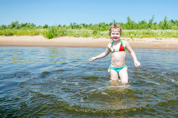 A cheerful happy girl is standing in the river and splashing water on a hot sunny day.