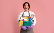 Happy young man holding bucket with cleaning supplies, pink studio background
