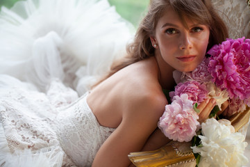 Bride in beautiful dress lying on sofa whit peonies in white studio interior. Romantic wedding style shot with sun light