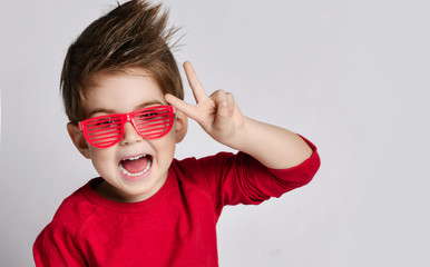 Little brunet child in sunglasses, red jumper. He opened mouth, showing victory sign, posing isolated on white background