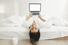 Happy Stylish Girl  Lying On Bed With Laptop With Blank Screen And Smiling In White Modern Room. Young Cheerful Woman Shopping Online Or Working Online From Home And Smiling. Freelance Mockup