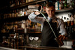 © fesenko - young barman professionally pours cocktail in shaker. Two glasses with ice stand on bar.