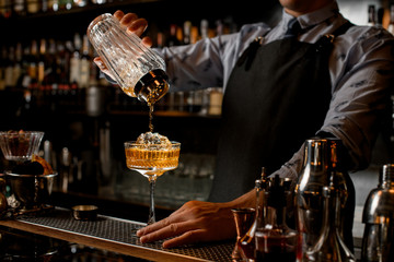 Professional bartender in black apron pours drink from shaker into glass.