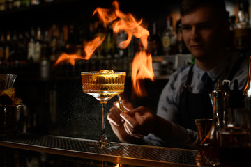 Young bartender carefully spray and makes fire over glass with cocktail