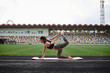© Natalia - Young brunette sportswoman, wearing black top and green leggings, doing yoga pose on city stadium on summer morning, Stretching training outside on pink yoga mat. Healthy active life concept.