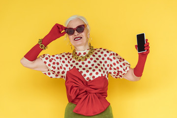 Wall Mural - Stylish senior woman smiling and holding smartphone with blank screen on yellow background