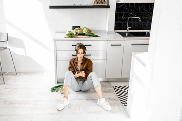 Wall Mural - Young woman relaxing on the kitchen floor, using mobile phone during the break while cooking at home