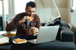 Young man eating waffle and drinking coffee while using laptop at home.