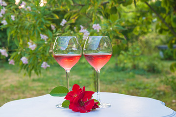 Romantic holiday composition. Two glasses of pink wine and red roses on white vintage table in the blooming garden