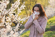 Young pretty woman sneezing in a park with blooming trees in spring. Pollen Allergy Symptoms