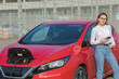© uflypro - Girl stands with phone near her red electric car and waits when vehicle will charged. Connecting the Charger Plug of an Electric Car.