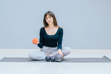 Sportive happy brunette woman trainer in sport clothing sitting on yoga mat showing balls pilates toning equipment in studio over grey wall background. Copy space