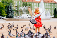Young Caucasian White Girl Feeding Many Pigeons, Some Flying Around Or Sitting Down. Blurred Fountain And Green Building In Background