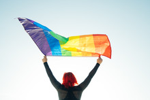 Woman Holding The Gay Rainbow Flag On Blue Sky Background. Happiness, Freedom And Love Concept For Same Sex Couples.