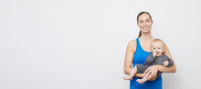 Portrait Of Happy Fit Mother And Her Cute Baby Looking Into The Camera, Stading On A Plain Background 