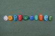 © yournameonstones - Copywriter, act or occupation of writing text for the purpose of advertising or other forms of marketing composed with multi colored stone letters over green sand
