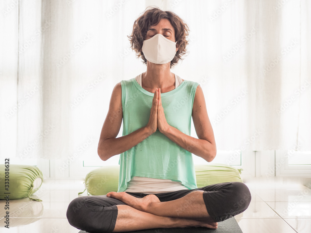 meditating woman wearing a medical face mask to protect from corona virus covid-19 in crossed legged lotus and praying namaste position on a yoga mat in a studio - obrazy, fototapety, plakaty 