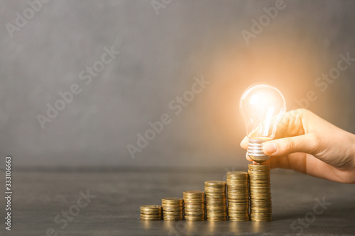 Female hand with glowing light bulb and coins on grey background. Concept of saving money