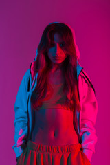 Wall Mural - Sexy young woman in a fashion top in a hoodie stands in the studio with a neon bright red light. Beautiful modern trendy girl fashion model with a slim body posing in a room with multi-colored color.