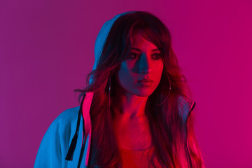 Wall Mural - Portrait of a beautiful young woman with long hair in a hoodie with a hood in a room with a bright pink neon light. Attractive girl in stylish clothes indoors with multi-colored color in disco style.
