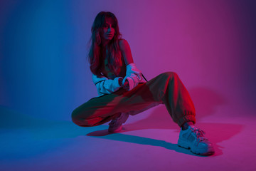 Wall Mural - Modern attractive young woman dancer in fashionable youth clothes in a white sneakers sits in a room with bright blue light. Sporty beautiful girl model poses in the studio with neon colorful color.