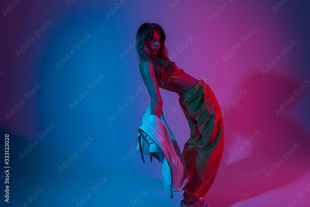 Attractive young woman dancer in fashionable youth clothes in a white sneakers dancing in a room with blue light.Sporty pretty beautiful girl model enjoys a dance in the studio with neon amazing color - obrazy, fototapety, plakaty 