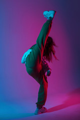 Wall Mural - Modern fashionable model young woman in stylish clothes in trendy gym shoes dancing indoors with bright multi-colored light. Sporty girl dancer posing in a room with bright neon pink-blue color. Style