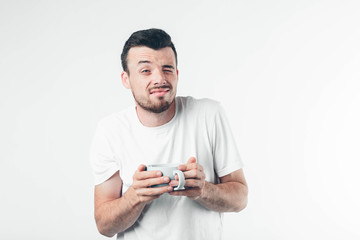 Young brunette man with beard dressed in white T-shirt. Guy looking at camera. One eye open second closed. He squints little. Holds white cup looks sleepy. Isolated over light background.