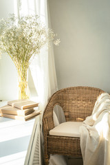 Cozy reading nook by the window with rattan wicker chair, delicate white gypsophila flowers, opened book and window light and shadows. Modern comfortable living room space. Slow living concept.